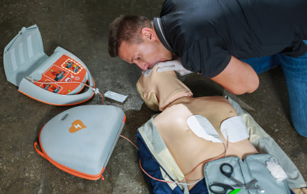 Man performing CPR on a training dummy next to a defibrillator